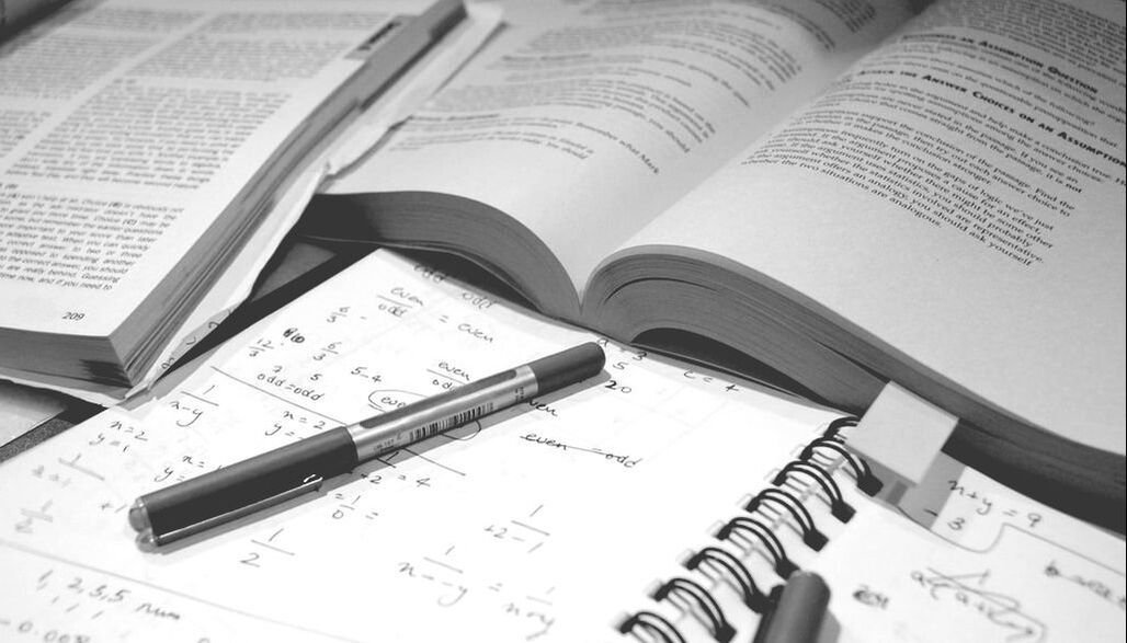 A black and white photograph of open textbooks, pens, and a notebook filled with mathematical equations.